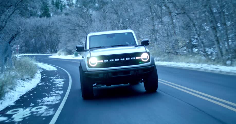 A white ford bronco with circular headlights drives along a winding road lined with snowdusted trees in a wintry landscape