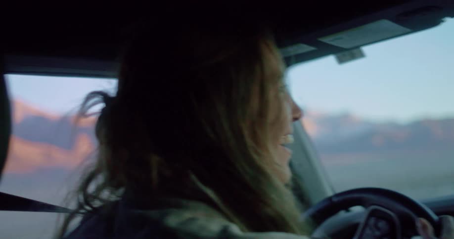 A woman smiles as she drives a vehicle with desert mountains visible through the window
