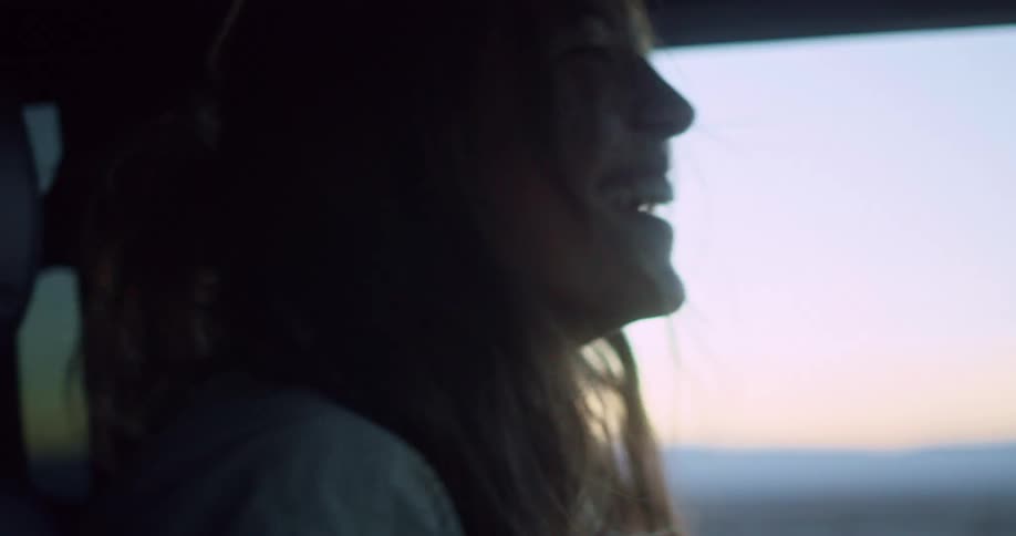 A woman with long hair smiles brightly while driving inside a vehicle during twilight