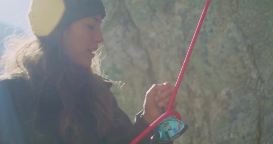 The video shows a closeup of a person who is rock climbing they are wearing a dark beanie and jacket and are seen pulling on a bright red climbing rope the background is a rocky surface and a bright light source creates a lens flare