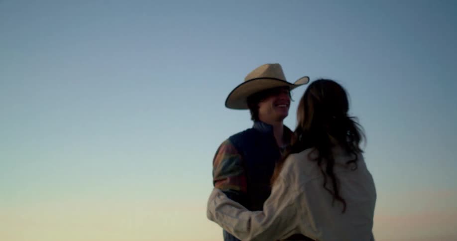 A happy couple stands close together at sunset in a wide open landscape with the man wearing a cowboy hat they playfully interact with the woman briefly taking the hat and putting it on his head as they smile