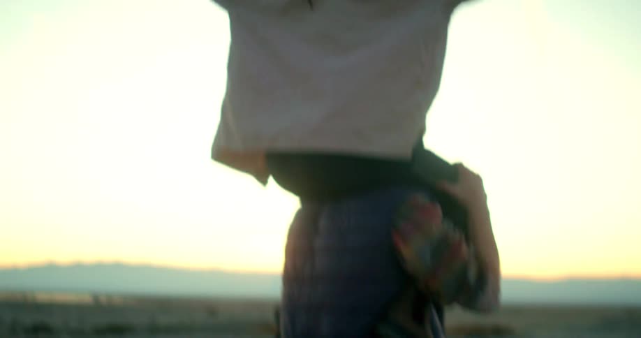 A person is giving a piggyback ride to another person in a desertlike landscape the person on top is raising their arms up and the one below is wearing a vest over a longsleeved shirt