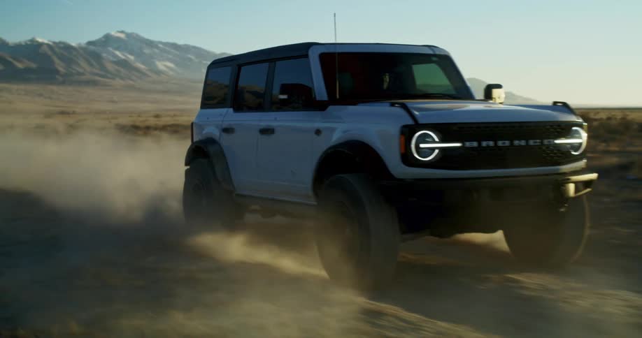 A white ford bronco kicks up dust while driving across a rugged desert plain with snowcapped mountains in the distance