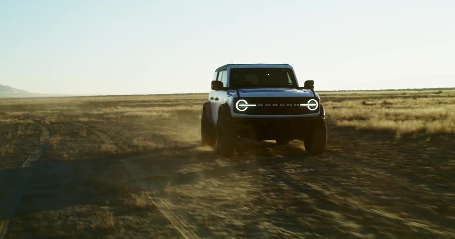 A white car driving away from the camera on a dusty dirt road through a wide open field with mountains in the distance