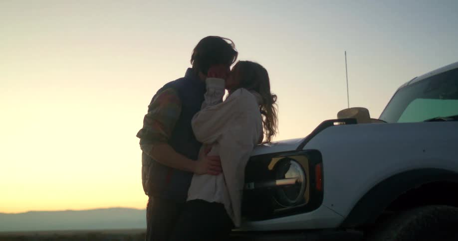 A loving couple hugs and shares a kiss in front of a white suv in a rustic open field at sunset