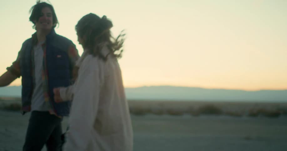 A man and woman couple are smiling holding hands as they walk and jump in a setting with the beach in the background and then they hug