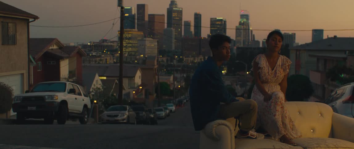 Woman and a man sitting on a white sofa in the middle of an urban street with skyscrapers of a metropolis in the background