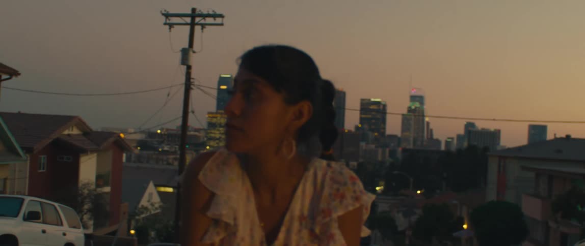 A couple sits on the back of a couch outdoors overlooking a cityscape at sunset the woman is wearing a floral dress and they are both looking at the city skyline as they talk