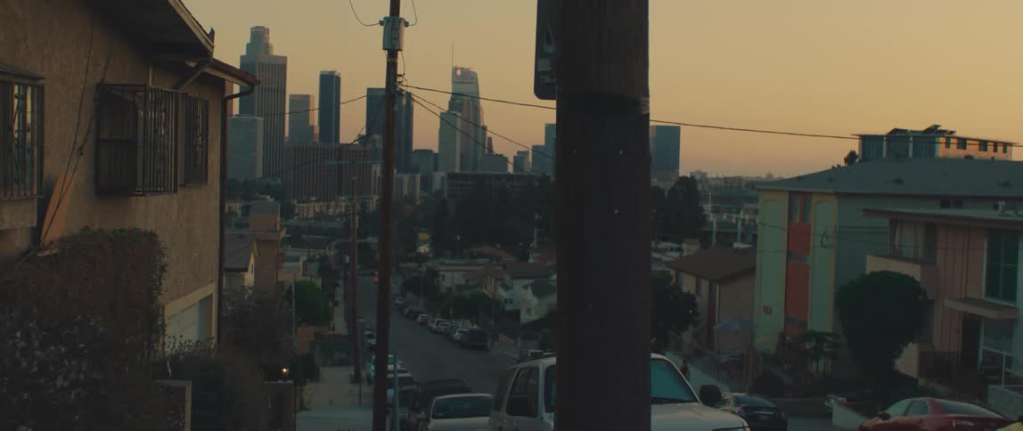 A neighborhood street in los angeles slopes downward toward the downtown skyline with tall buildings silhouetted against the warm glow of sunset