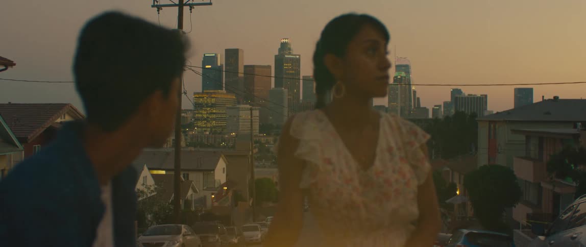 The video shows two people a man and a woman standing outside during the evening with a city skyline in the background the woman in a white floral dress is looking off into the distance the man with his back mostly to the camera is looking at her