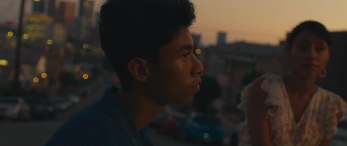 Two teenagers a girl and a boy stand on a rooftop with the cityscape in the background