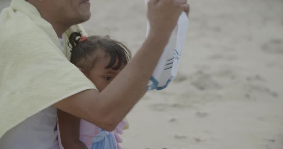 A toddler sits on a man's lap on a sandy beach while he folds a towel