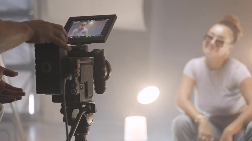 The video shows a film shoot in progress with the focus on a professional camera rig in the foreground hands are seen adjusting the cameras settings and a monitor attached to the camera shows a woman being filmed the woman being filmed is in the bac