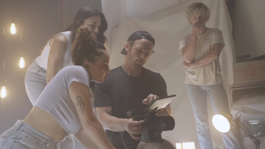The video shows a film crew gathered around a camera and a tablet likely reviewing footage or planning a shot a man in a black tshirt and a backward baseball cap is holding the camera and looking at the tablet two women and a man are standing around h