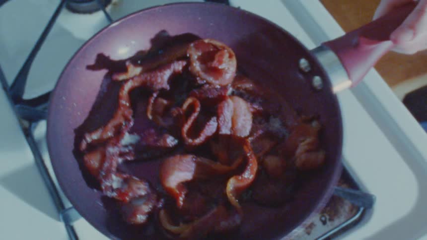 Several strips of bacon sizzling and cooking in a darkcolored frying pan on a gas stovetop