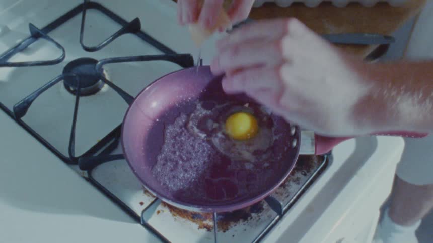 Hands cracking an egg into a purple frying pan on a lit stove
