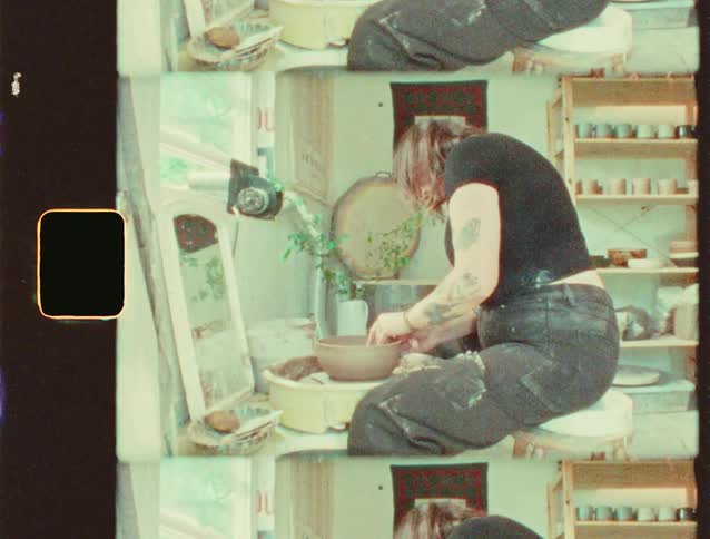 A potter is sitting at a pottery wheel and working on a clay pot the person has tattoos on their arms and is in a room with shelves full of ceramics