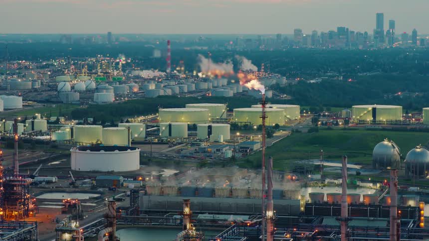 An expansive industrial oil refinery with storage tanks and smokestacks emits flames and smoke against a distant city skyline at dusk