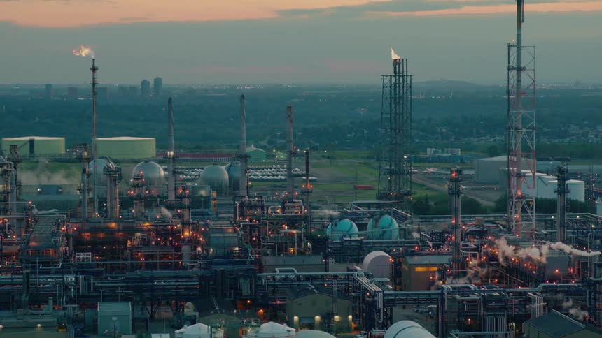 A sprawling oil refinery complex at dusk with flare stacks and storage tanks