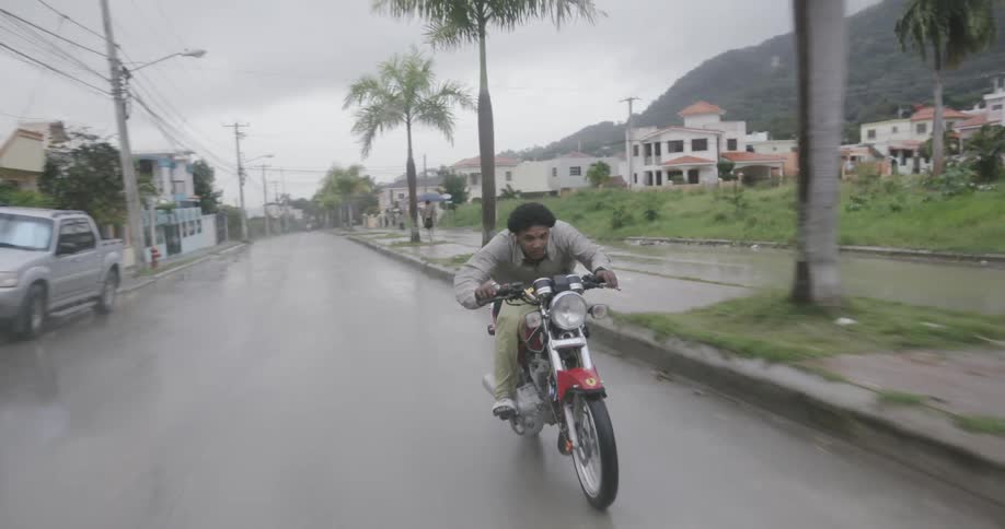A man rides a motorcycle through a town while it rains