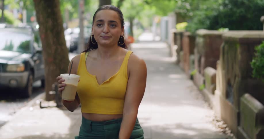 A woman walks down a city sidewalk drinking iced coffee