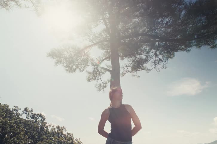 Woman standing under a tree looks to the side with the sun shining in the background