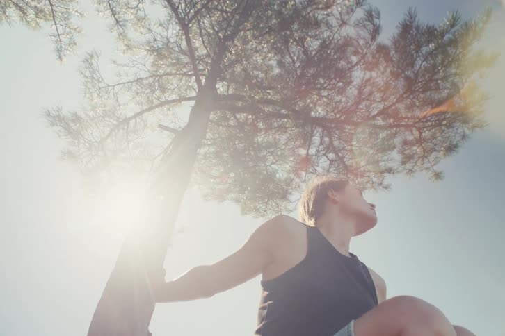 From a low angle a woman resting under a tall pine tree on a sunny day turns to look directly at the camera after a bright lens flare passes