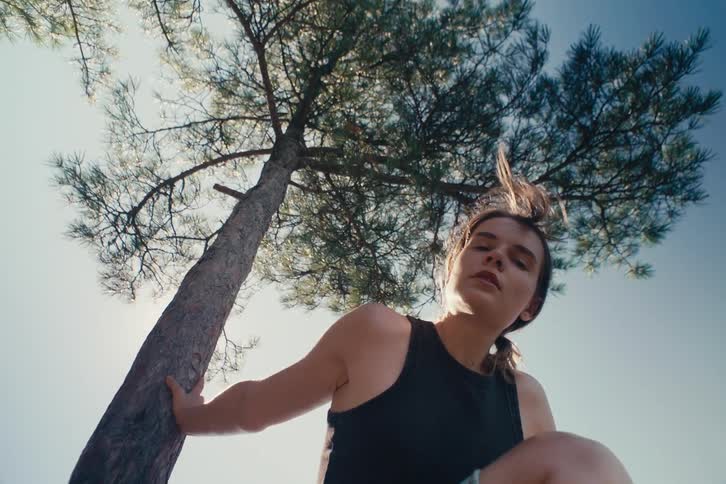 Woman leans against a pine tree while crouching outside on a sunny summer day