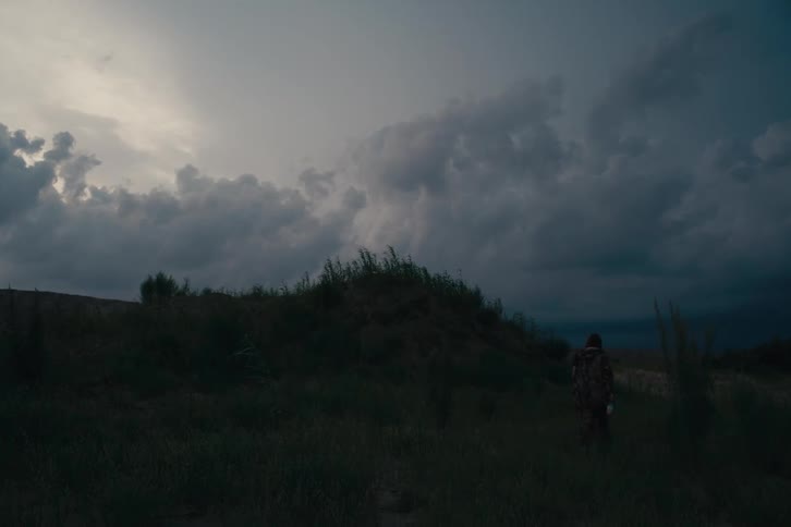 A person walks through a grassy field at dusk beneath a dramatic sky filled with dark rolling clouds