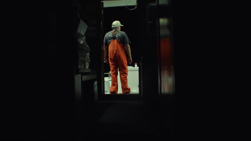 A man in orange overalls and a cap stands on a brightly lit deck at night viewed from a dark doorway
