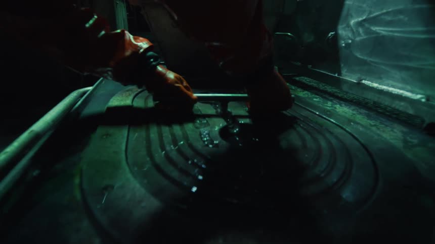 A person in orange gloves working on a greasy dark machine in an industrial environment