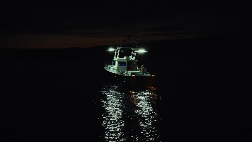 A boat floats on a lake at night with a person moving around inside as the boats light illuminates the surroundings