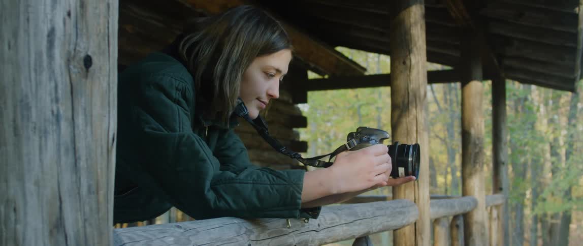 Female photographer leans on a porch rail while taking a picture at a cabin in the woods on an autumn day
