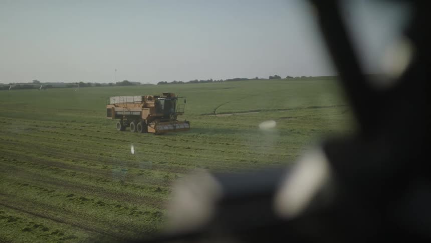 A large combine harvester is visible through a blurred foreground actively working in a wide flat green field under a clear sky