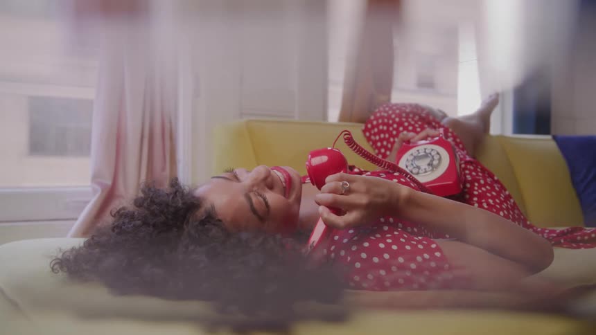 A video shows a young woman with curly dark hair wearing a red polkadot twopiece outfit she is lying on a yellow couch and holding a red rotary telephone to her ear she appears to be laughing