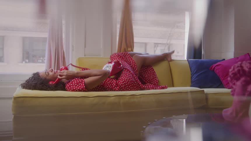 A video shows a young woman with curly hair lying on a yellow couch she is wearing a red polkadot twopiece outfit and is talking on a red rotary telephone smiling and laughing