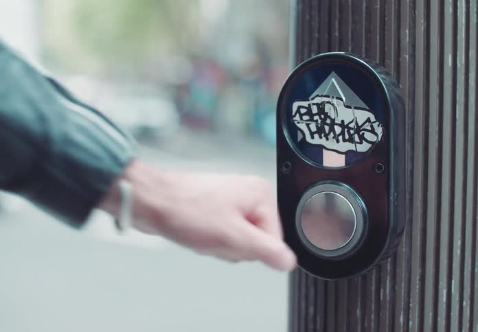 A video shows a closeup shot of a walk signal button covered in graffiti on a city street a persons hand reaches out and presses the button a white truck is out of focus in the background
