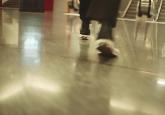 A video shows a lowangle shot of a persons feet walking on a shiny floor the person is wearing black pants and black converse sneakers with white soles the background is blurred but an escalator is visible