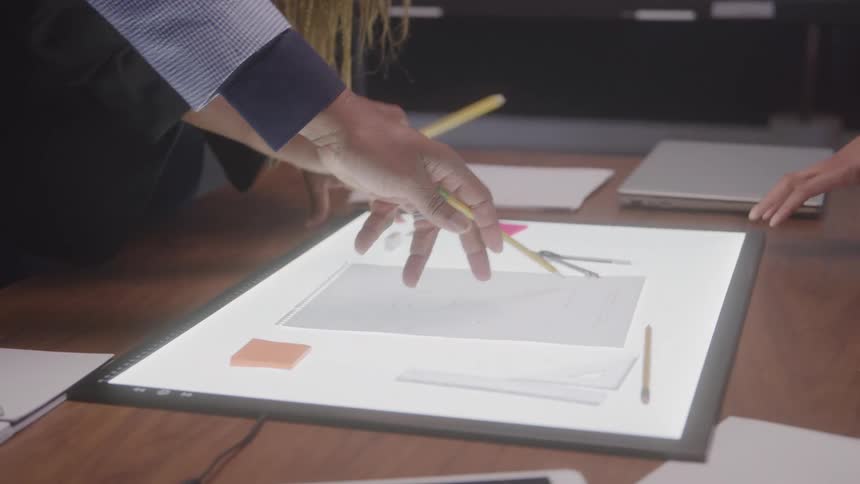 A video shows a closeup shot of two peoples hands working together on a light board on a desk they appear to be discussing a design or drawing with pencils a ruler and a compass nearby