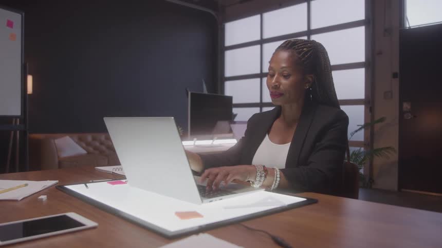 A video shows an african american woman with braided hair sitting at a large wooden desk in an office she is looking at her laptop and typing and there are papers pens and a smartphone on the desk in front of her she is wearing a black blazer