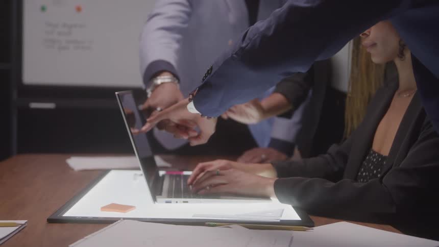 Several people in business attire are gathered around a laptop with multiple hands pointing at the screen