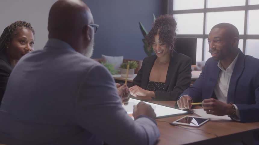 Four business professionals three men and one woman are seated around a conference table in an office actively engaged in a discussion with one woman pointing at a glowing surface on the table