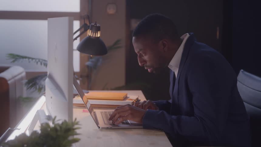 A black man works with a laptop while sitting at a desk in an office at night