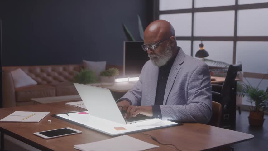 A distinguished older man with a grey beard and glasses types on a laptop at a wooden desk in an office with a bright light pad beneath the laptop and a cozy seating area in the background