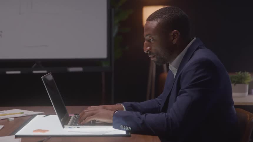 Man in a suit typing on a laptop at a lit desk in a modern office
