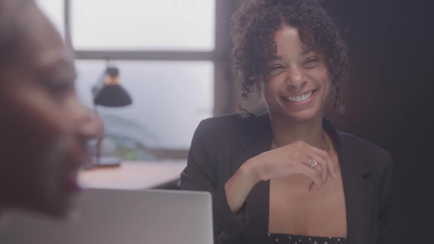 A woman in business attire with curly hair smiles warmly appearing engaged and cheerful during a discussion