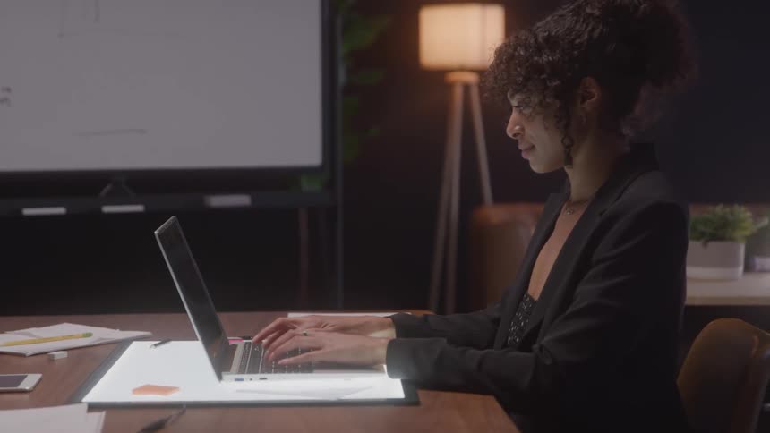 Woman in a black blazer typing on a laptop in an office room lit by a lamp