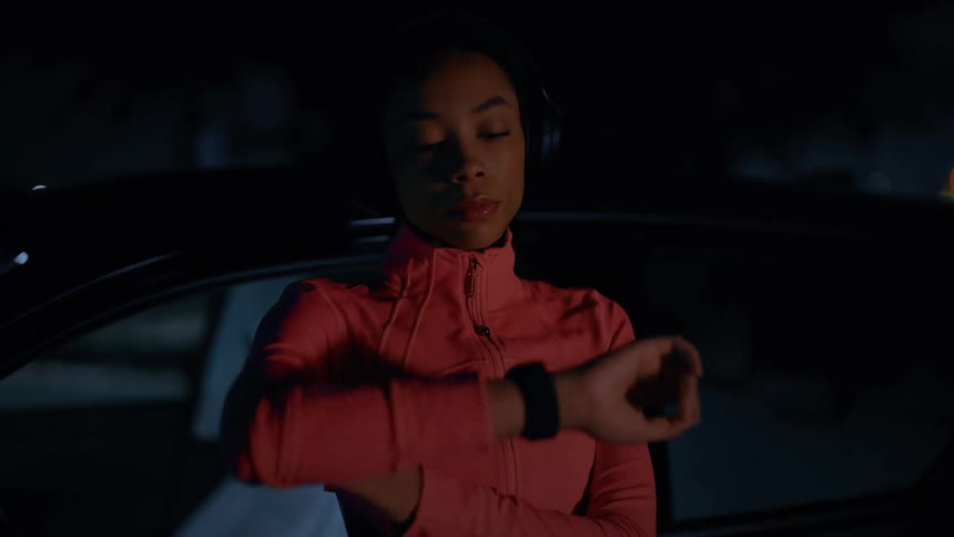 Close up of a young black woman wearing her headphones and checking her wristwatch before continuing jogging outdoors at night