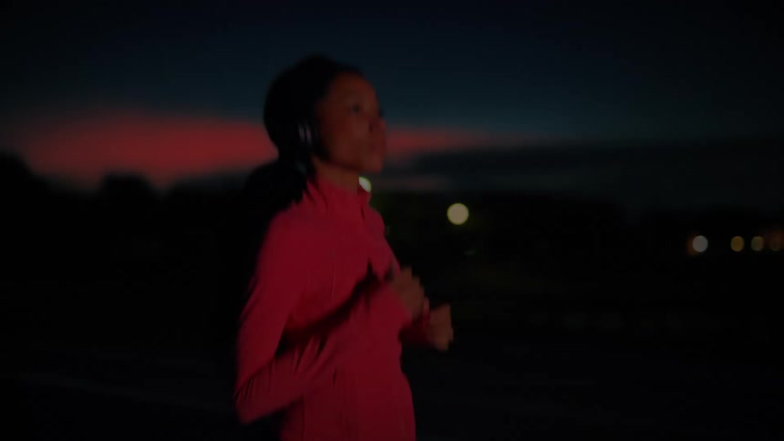 A young black woman wearing headphones jogs outdoors on a road at night
