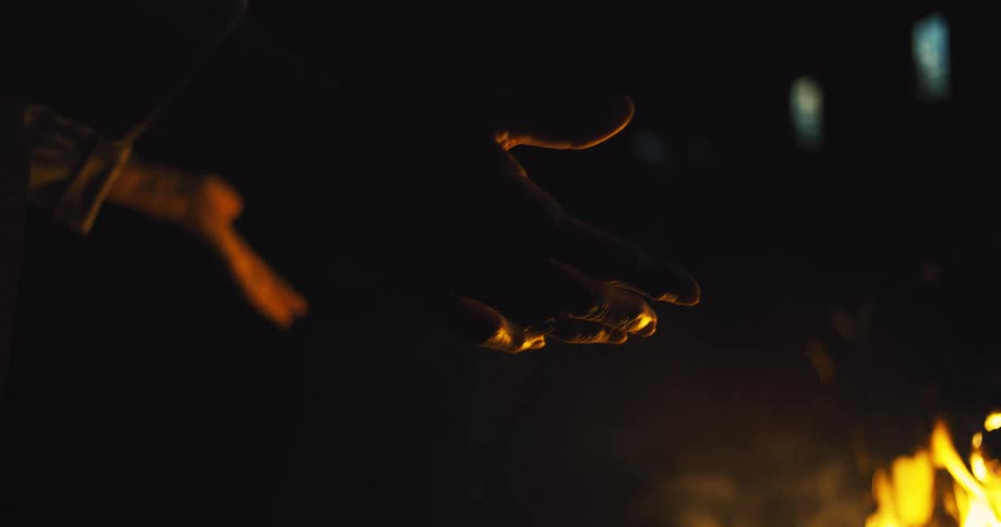 A persons hand backlit by the flickering glow of a fire as they hold their hands out to warm themselves